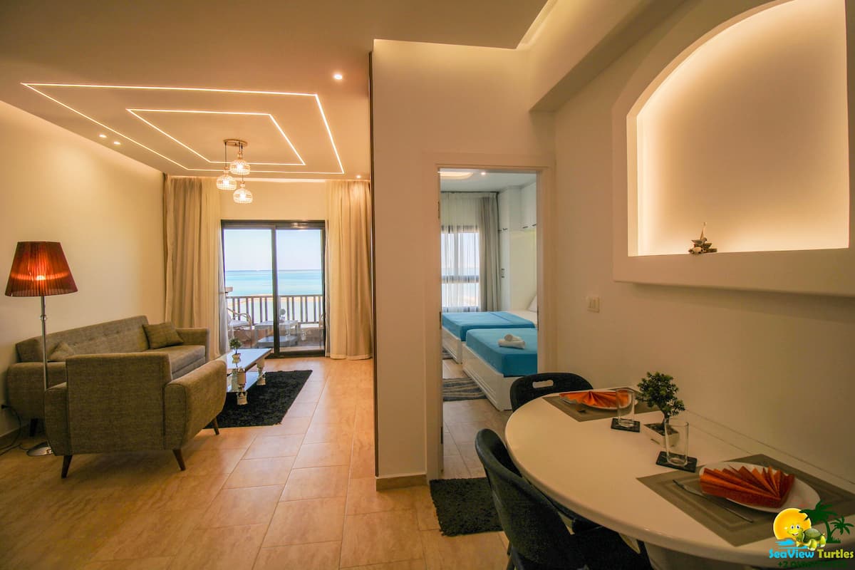 A modern hotel room with a sofa, a dining table, and a view of the sea through a glass door.