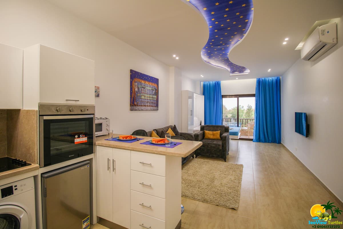 A modern kitchen with a built-in oven and microwave, a dining area with a table and chairs, and a living room with a sofa and a TV.