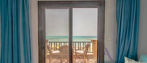 Una habitación con puerta corrediza de vidrio que da a un balcón con vistas al océano. Hay sillas de mimbre y una mesita en el balcón.