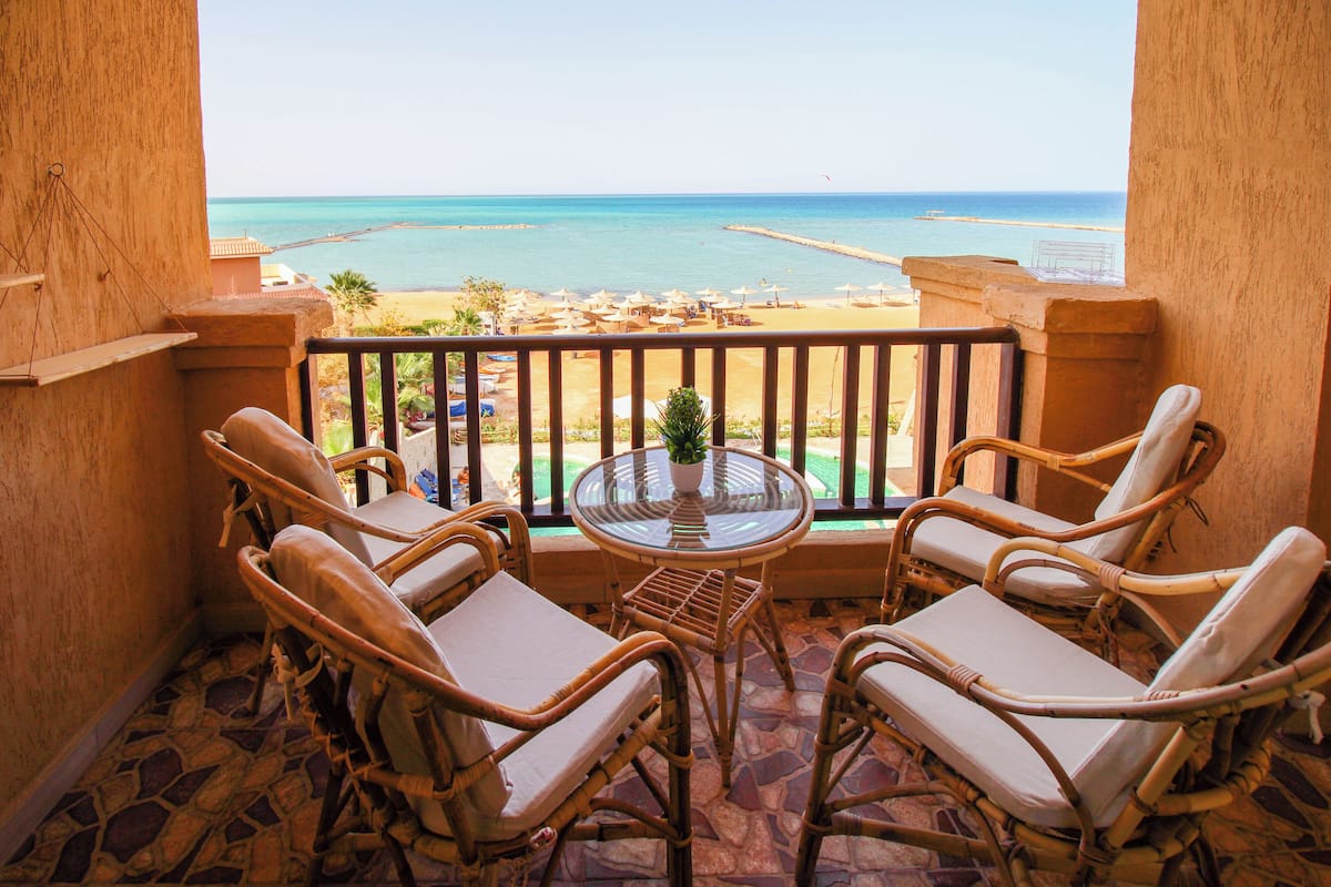 A balcony with wicker chairs and a glass table, overlooking a beach and the sea.