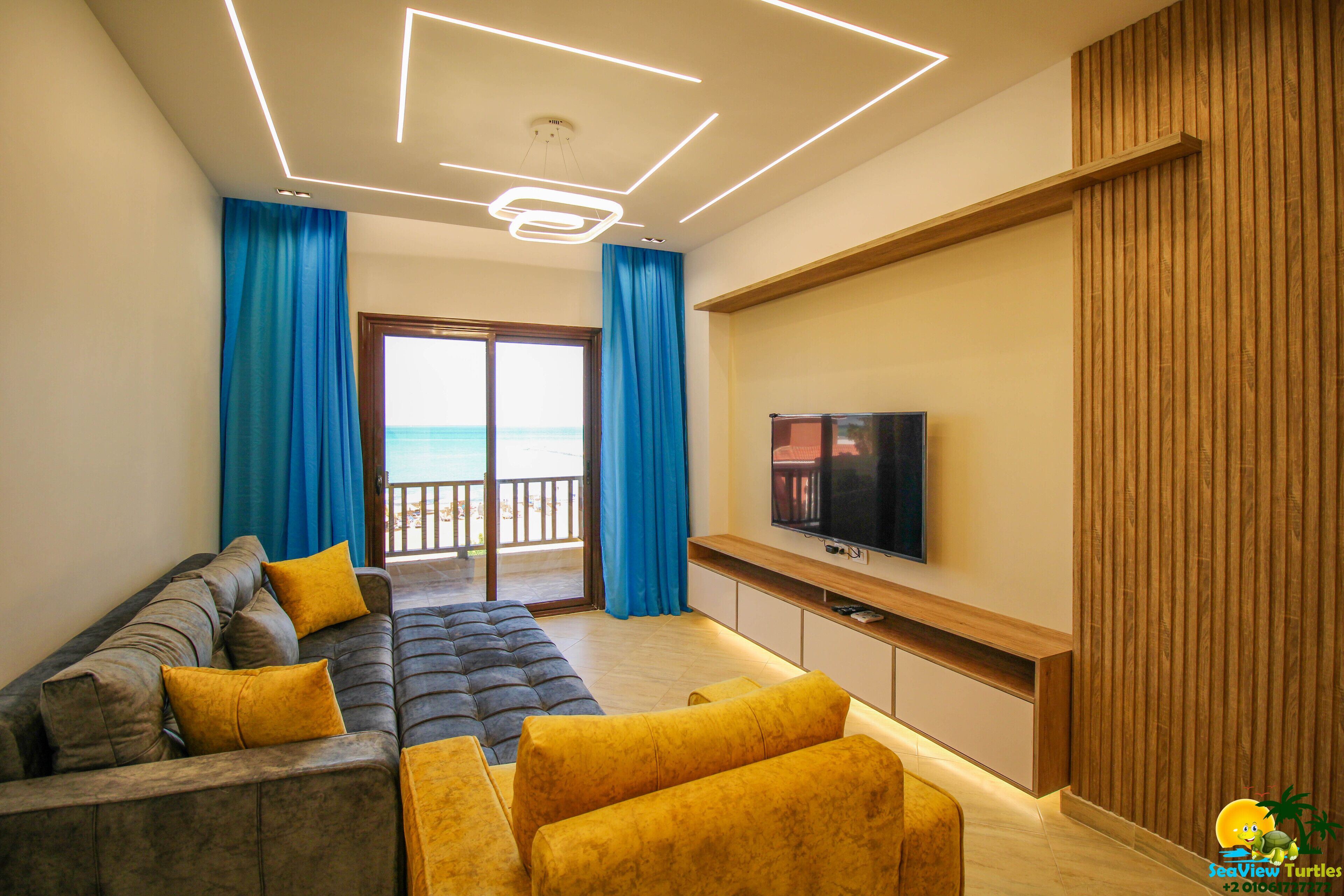 Een moderne woonkamer met een bank, een gele poef en een flatscreen televisie aan de muur.