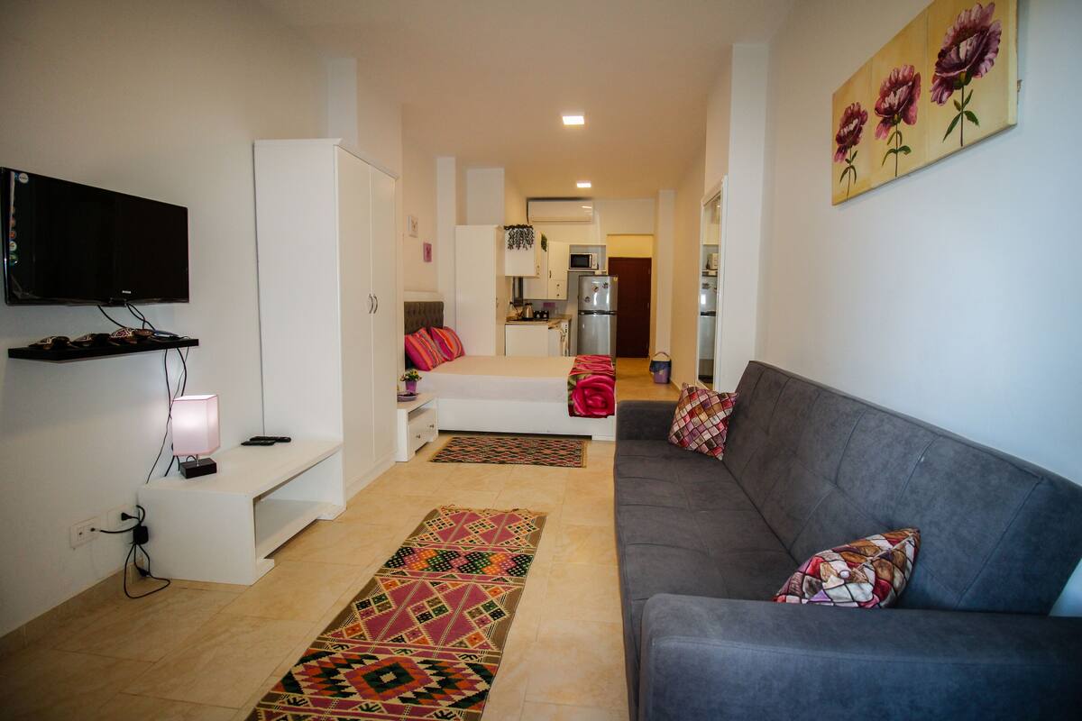 A modern living room with a grey sofa, a flat-screen TV mounted on the wall, and a kitchen in the background.
