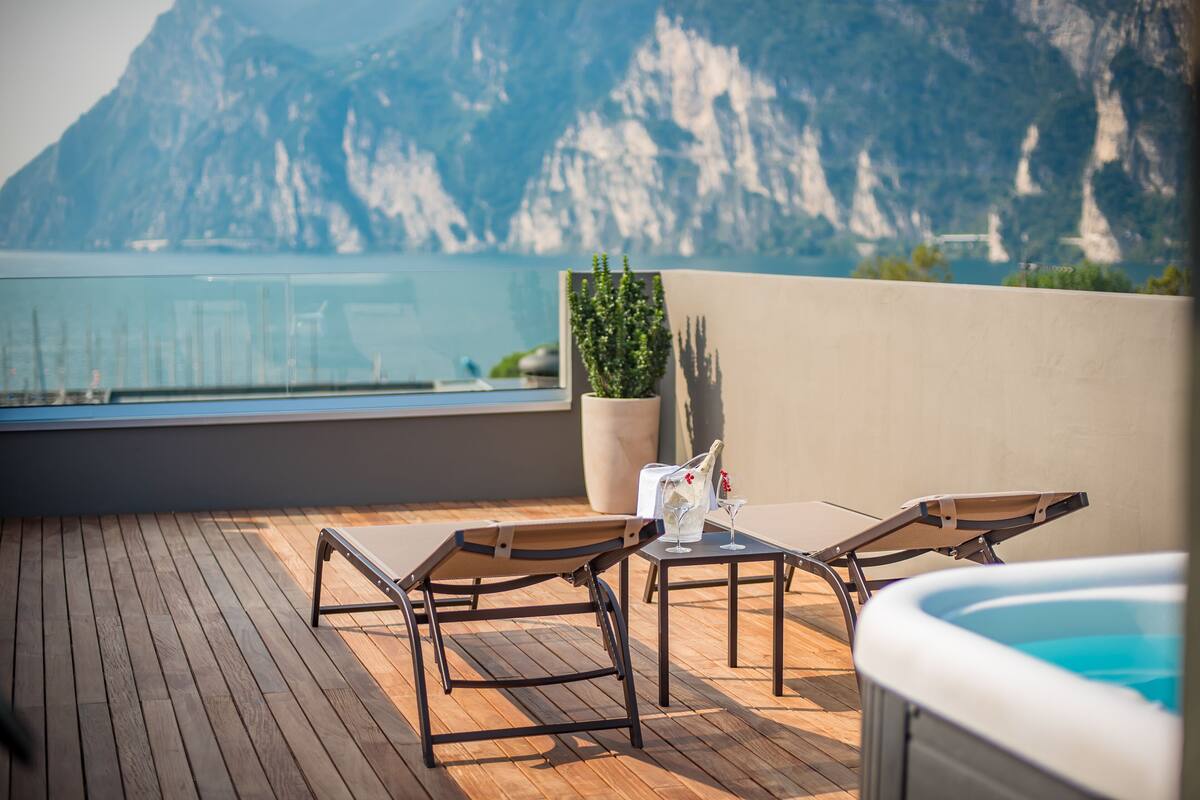 Una terraza de madera con sillones de descanso y un jacuzzi, con vista a un cuerpo de agua y montañas.