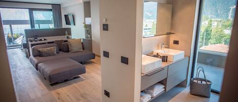 A modern hotel room with a large bed, a sofa, a desk, and a bathroom with a large mirror and sink.