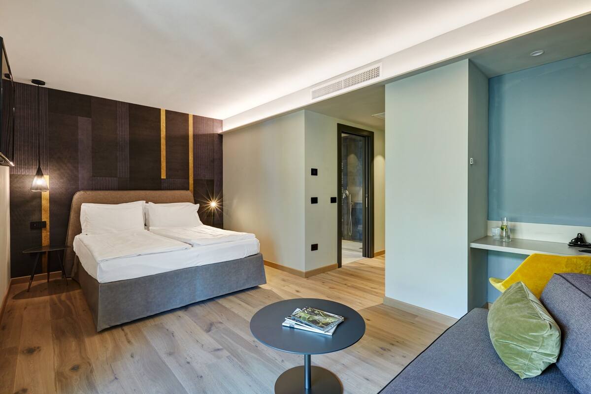 Una habitación de hotel moderna con una cama grande, una pequeña mesa redonda con revistas y una zona de estar con cojines.
