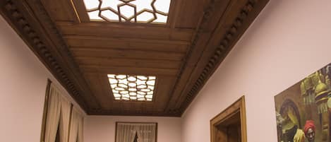 A room with wooden flooring, a wooden ceiling with a geometric skylight, a wooden door, a framed painting on the wall, and a chair with a potted plant.