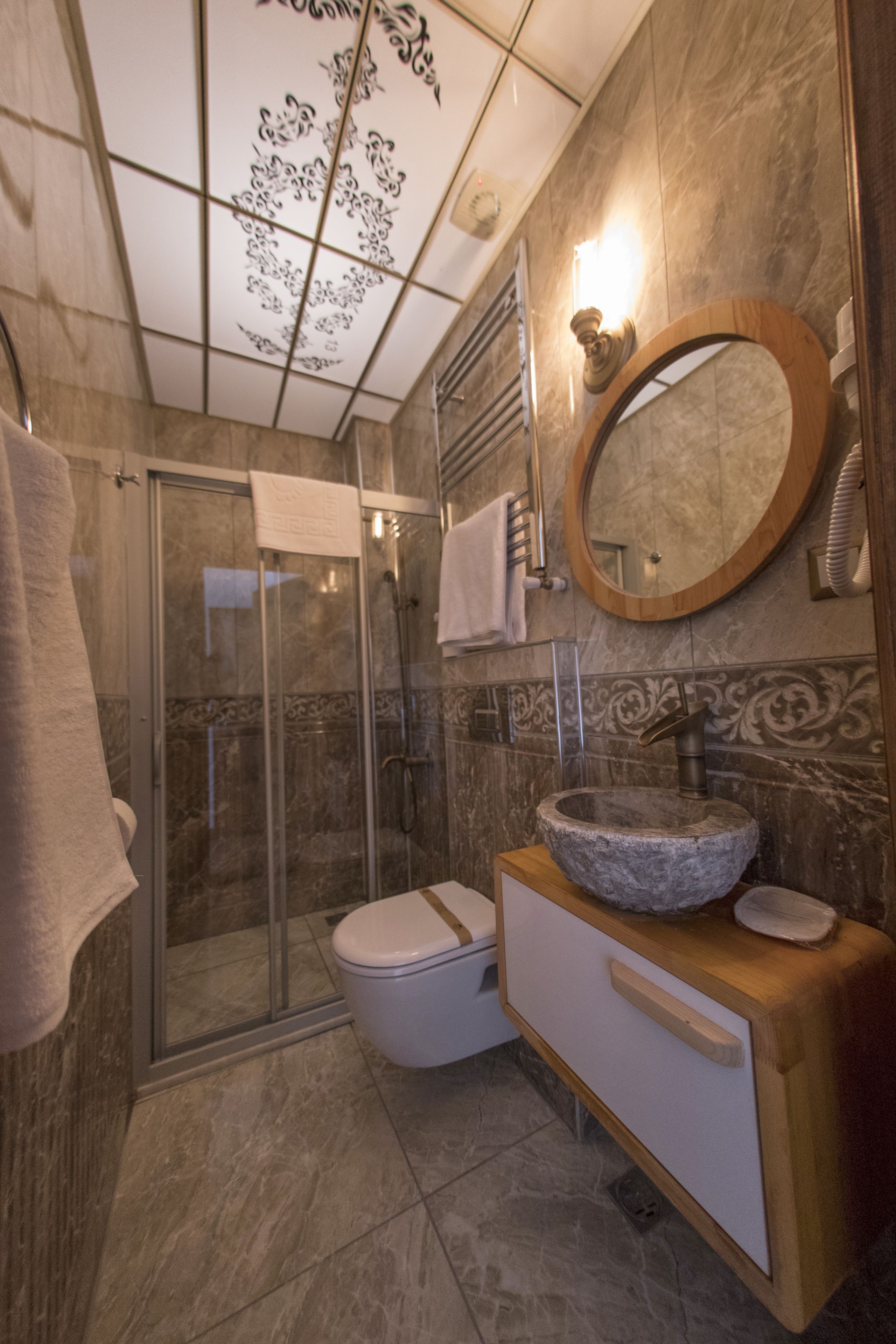 Un baño con cabina de ducha de vidrio, un inodoro blanco, un tocador de madera con lavabo de piedra y un espejo redondo.