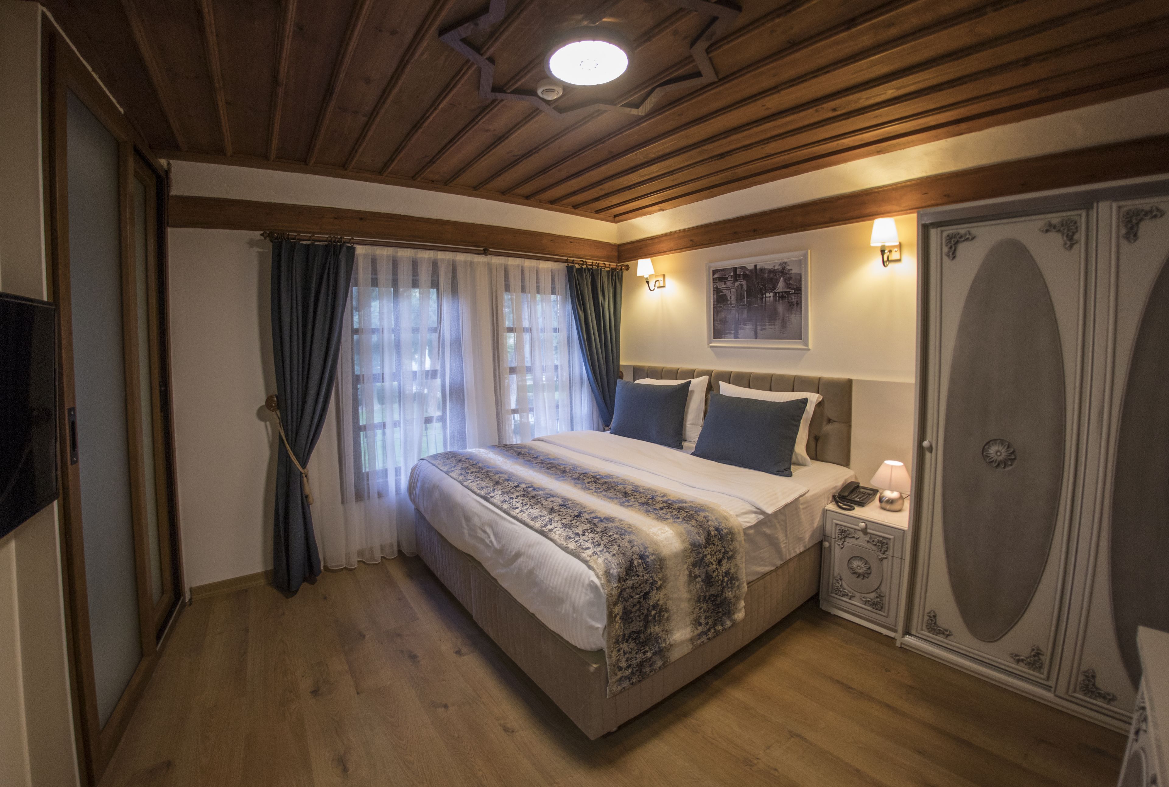 A bedroom with a wooden ceiling, a bed with a patterned blanket, a bedside table with a lamp, and a wardrobe.