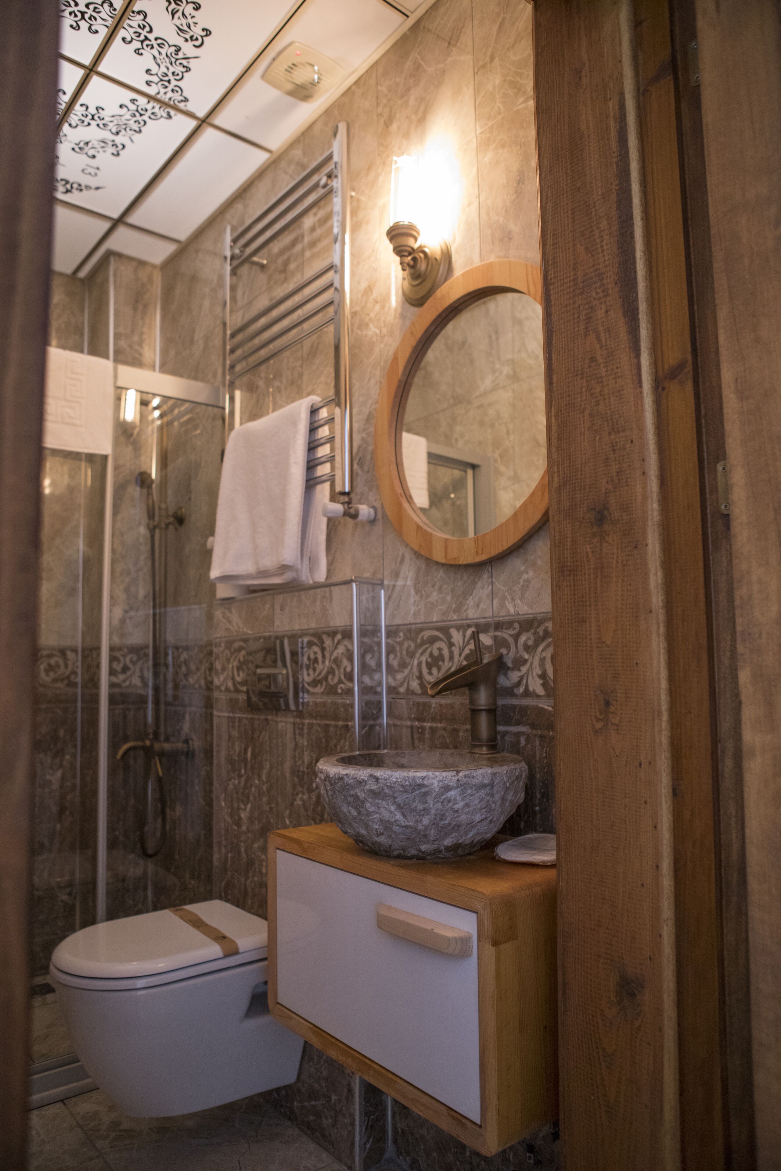A bathroom with a marble wall, a wooden vanity, a white toilet, a glass shower enclosure, and a mirror.