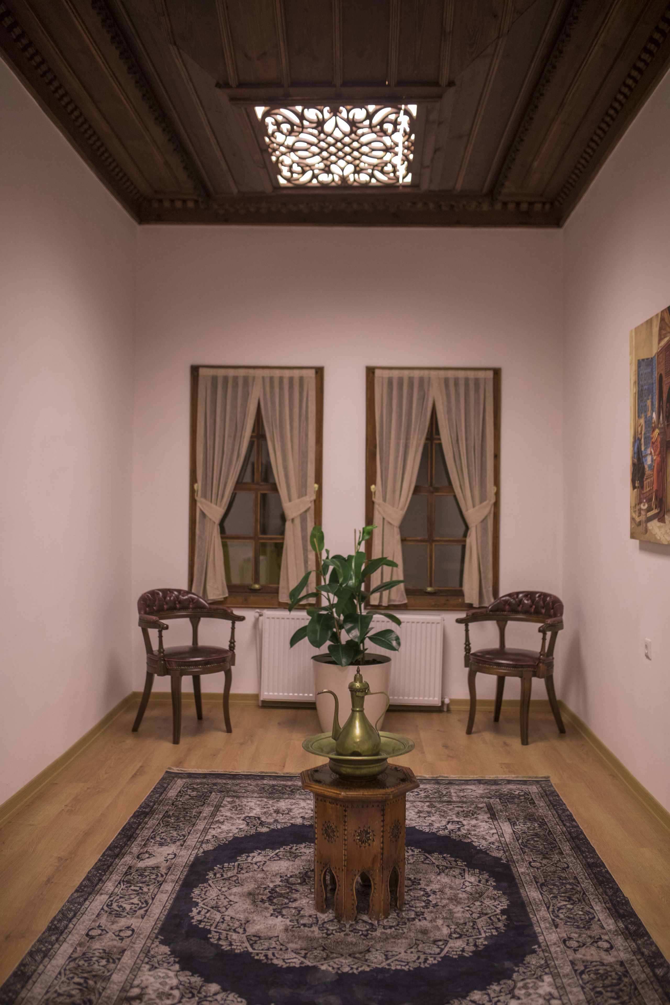 Una habitación con dos ventanas, un radiador, techo de madera y una alfombra estampada.