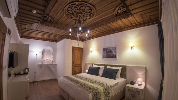 A bedroom with a wooden ceiling, a bed with a green blanket, bedside lamps, and a nightstand with a lamp. - Bayezid Han Konak (Amasya)
