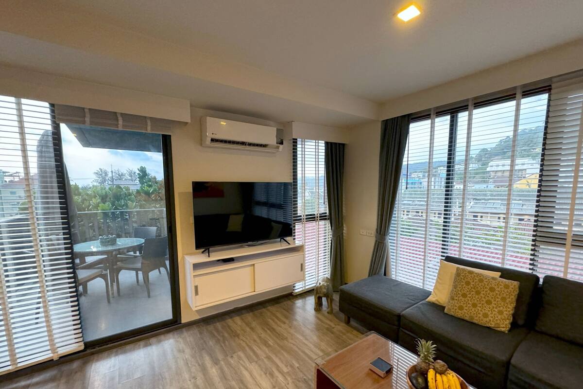 A modern living room with a flat-screen TV, a sofa, a coffee table with fruits, and a balcony with a view of the city.