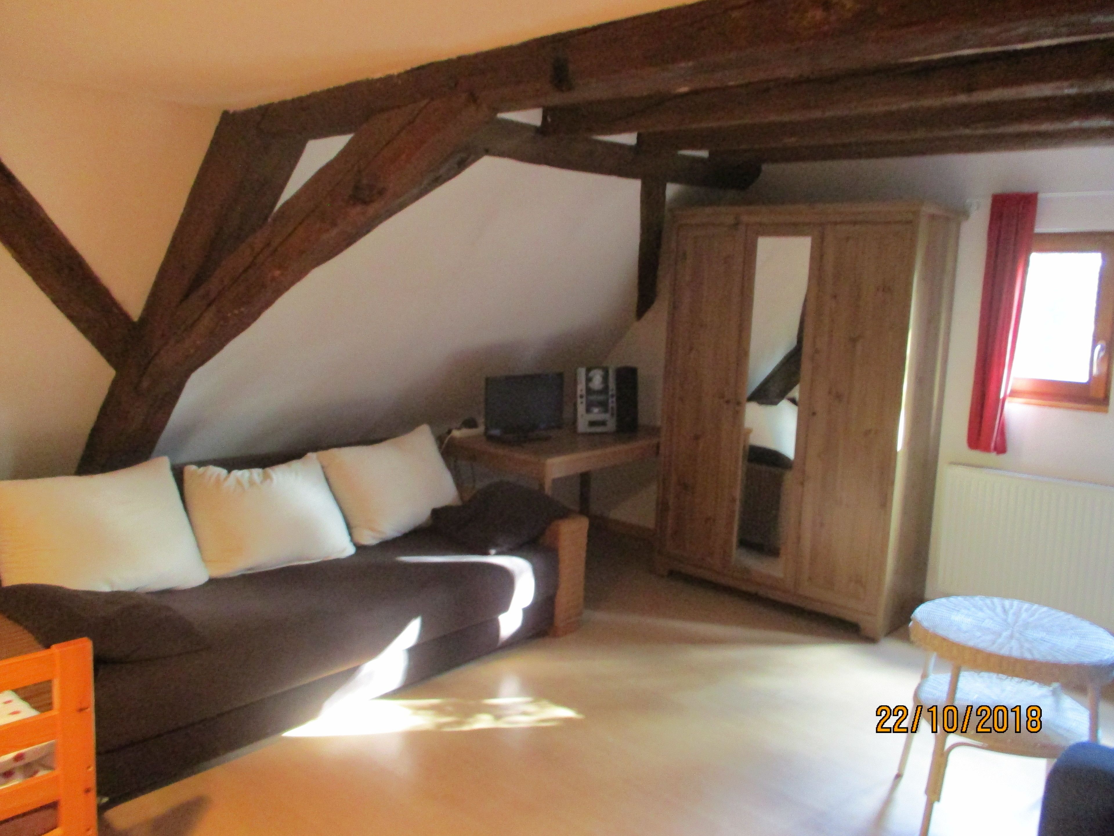 An attic bedroom with a sloped ceiling, a wooden beam structure, a sofa with cushions, a small table with a television, a wardrobe, and a window with a red curtain.