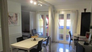 A dining area with a white table, black chairs, and a small table lamp. There is a large window with curtains, and a door leading to an outdoor area.