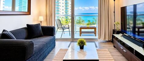 A modern living room with a sofa, coffee table, TV, and a balcony with a view of the ocean and buildings.
