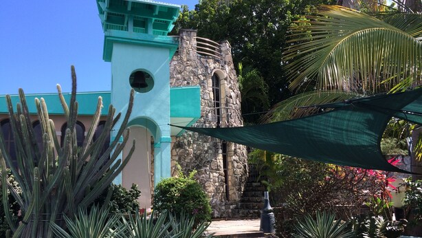 A building with a stone tower and a bell tower, surrounded by cacti and tropical plants.