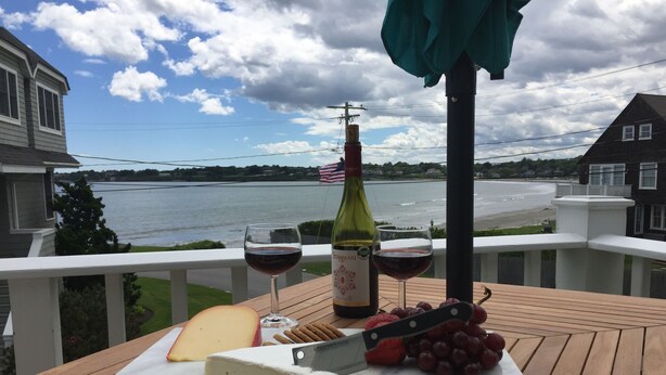 Una terraza de madera con una tabla de mármol que sostiene un trozo de queso, un cuchillo, una botella de vino y una copa de vino tinto.