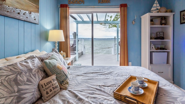 A bedroom with a bed, a tray with cups, a bookshelf, and a view of the water.