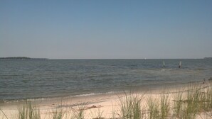 A beach with sand and grass in the foreground, a calm sea, and a distant landmass.