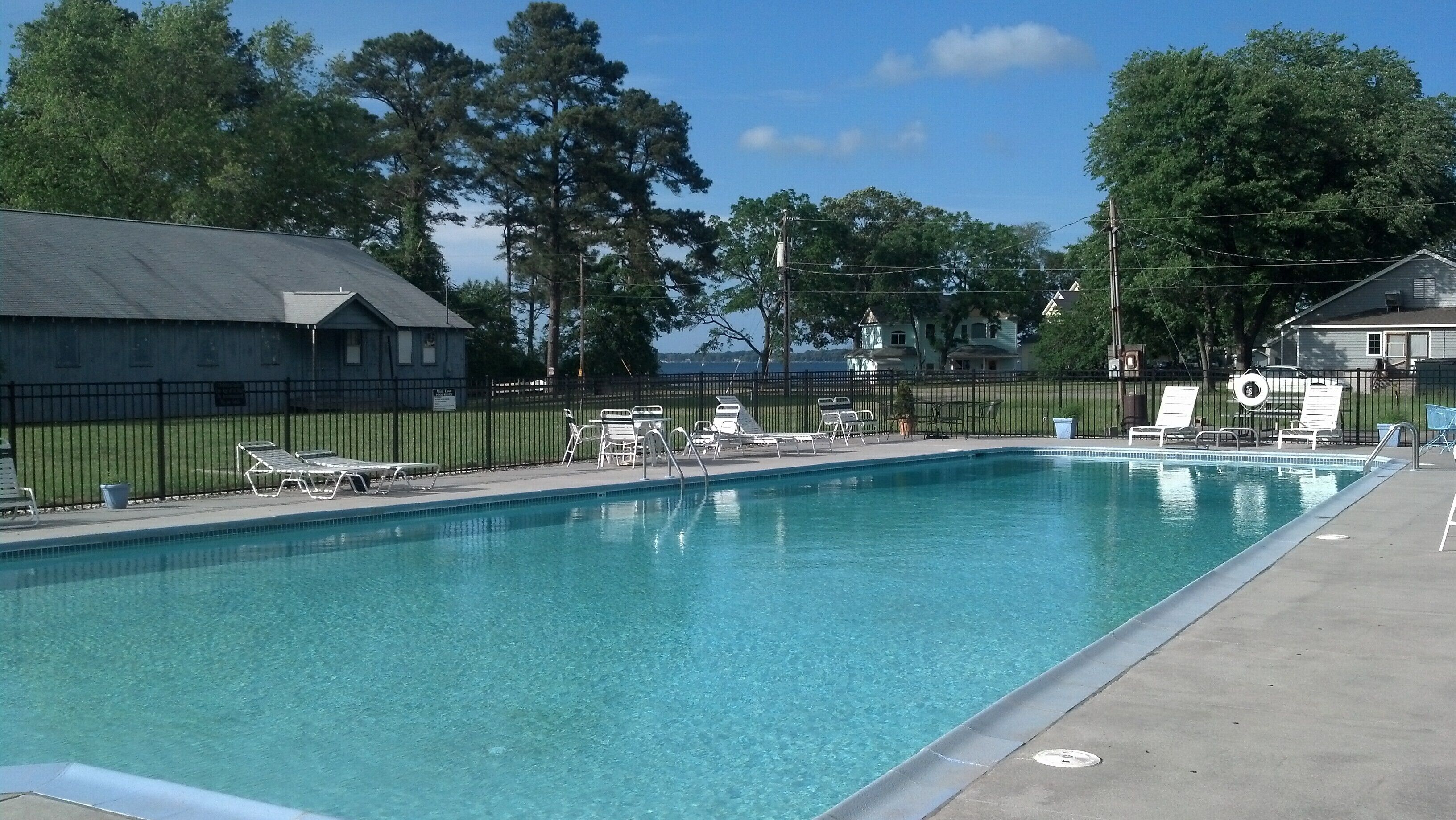 A rectangular swimming pool with clear blue water, surrounded by a black metal fence. There are several white lounge chairs and a small lifeguard stand. The pool area is adjacent to a paved walkway, and there are trees and buildings in the background.