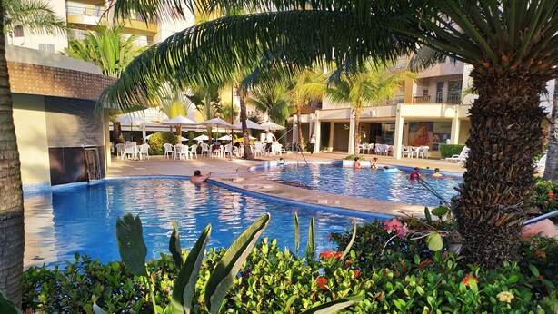 Piscina rodeada por vegetação exuberante e palmeiras, com área de estar ao ar livre e um edifício ao fundo.