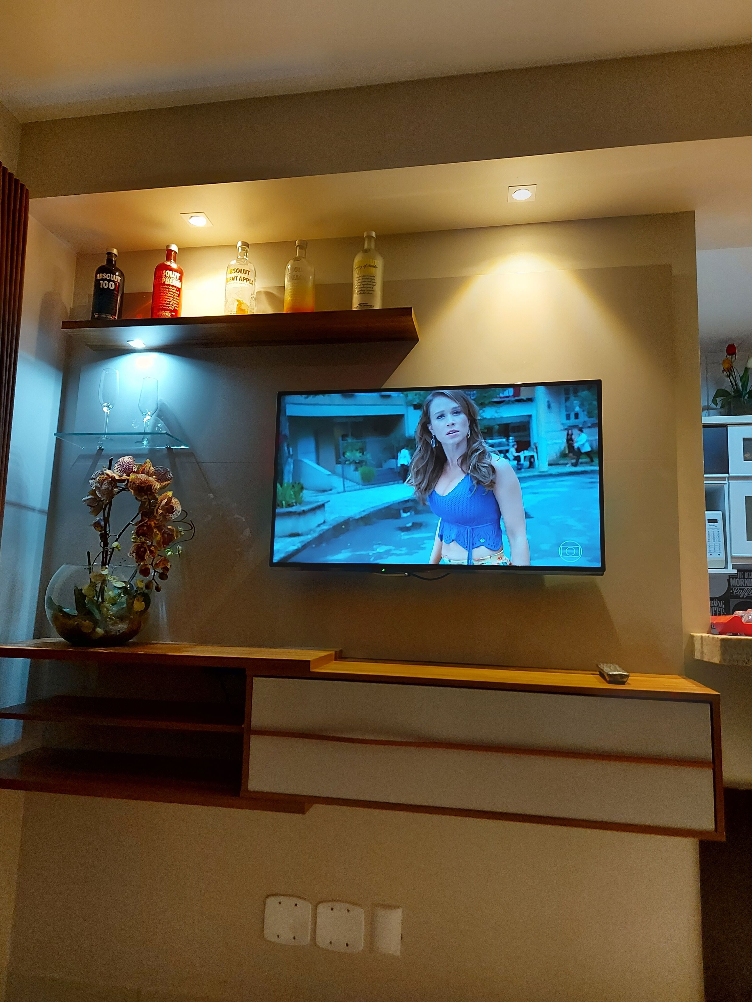 A television mounted on a wall with a shelf above it displaying bottles and a vase with flowers.