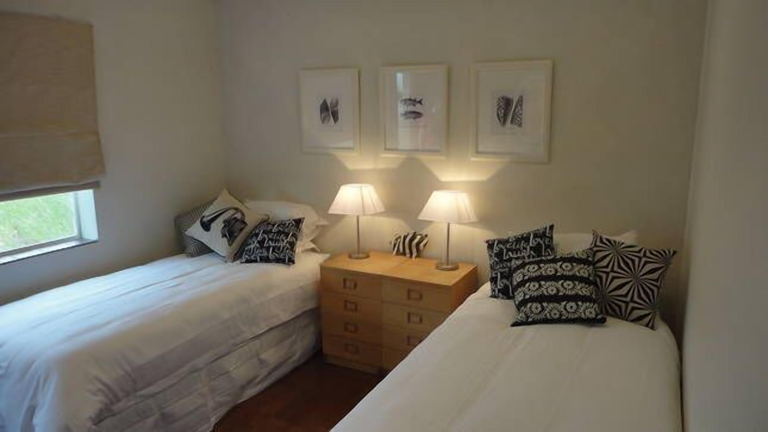 A bedroom with two single beds, a nightstand with two lamps, and three framed pictures on the wall.