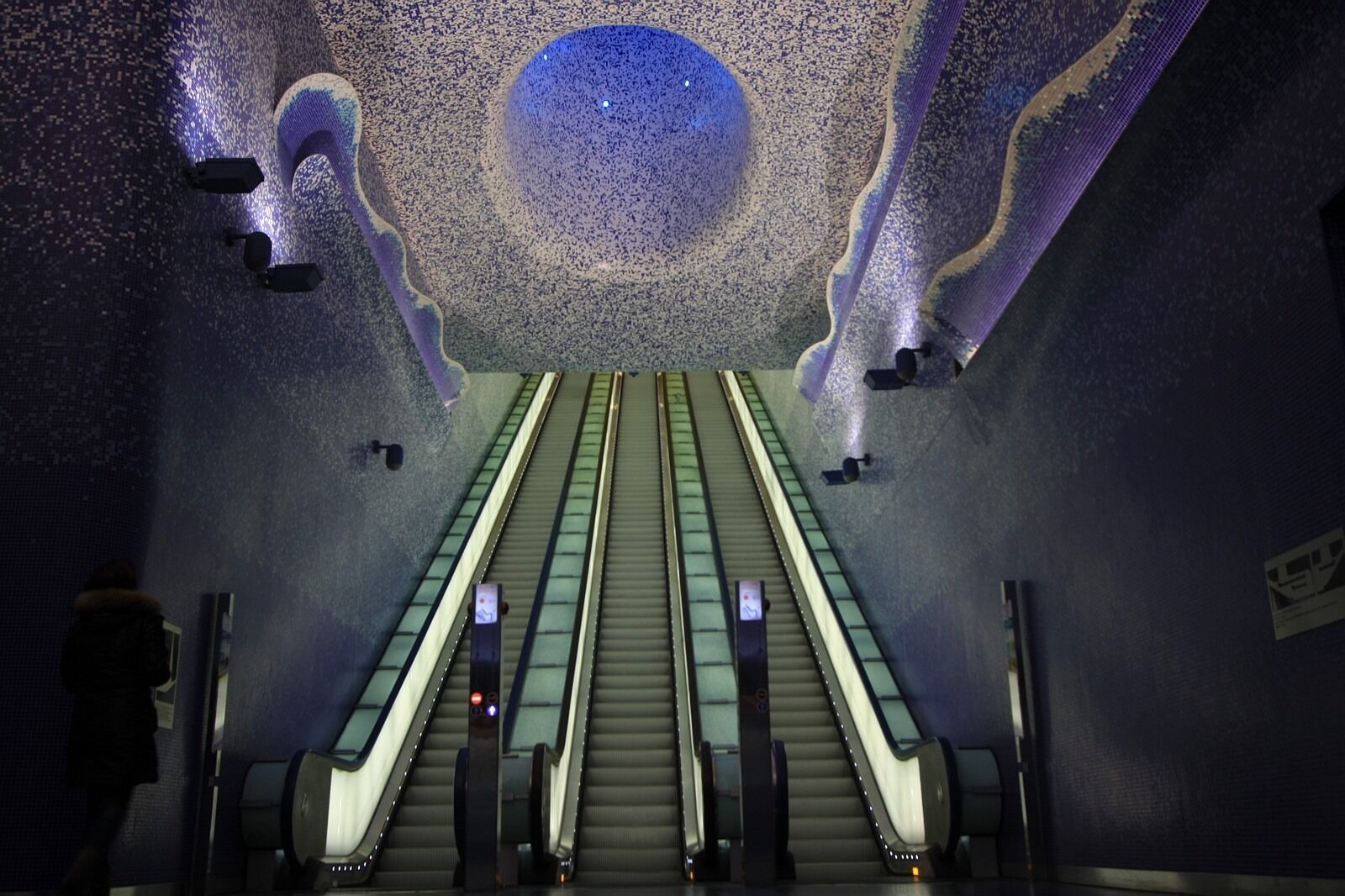 Eine Rolltreppe in einem Innenraum mit einer Decke, die ein großes, kreisförmiges, beleuchtetes Design zeigt.