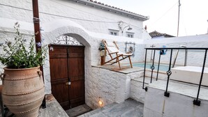 A white building with a stone door, a wooden chair by a pool, and a large terracotta pot. - Villa Andreas (Hydra)
