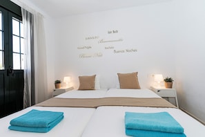 A double bed with two blue towels, a bedside table with a lamp and a plant, and a wall with multilingual "Good Night" signs.