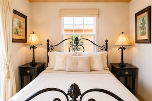 Un dormitorio con una cama, dos mesitas de noche, dos lámparas, una ventana con vistas y dos cuadros en las paredes.