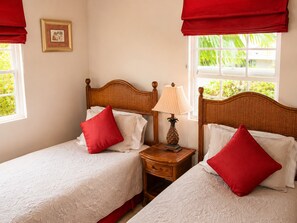 A double bed with red pillows and a nightstand with a lamp, in a room with a window and a framed picture on the wall.