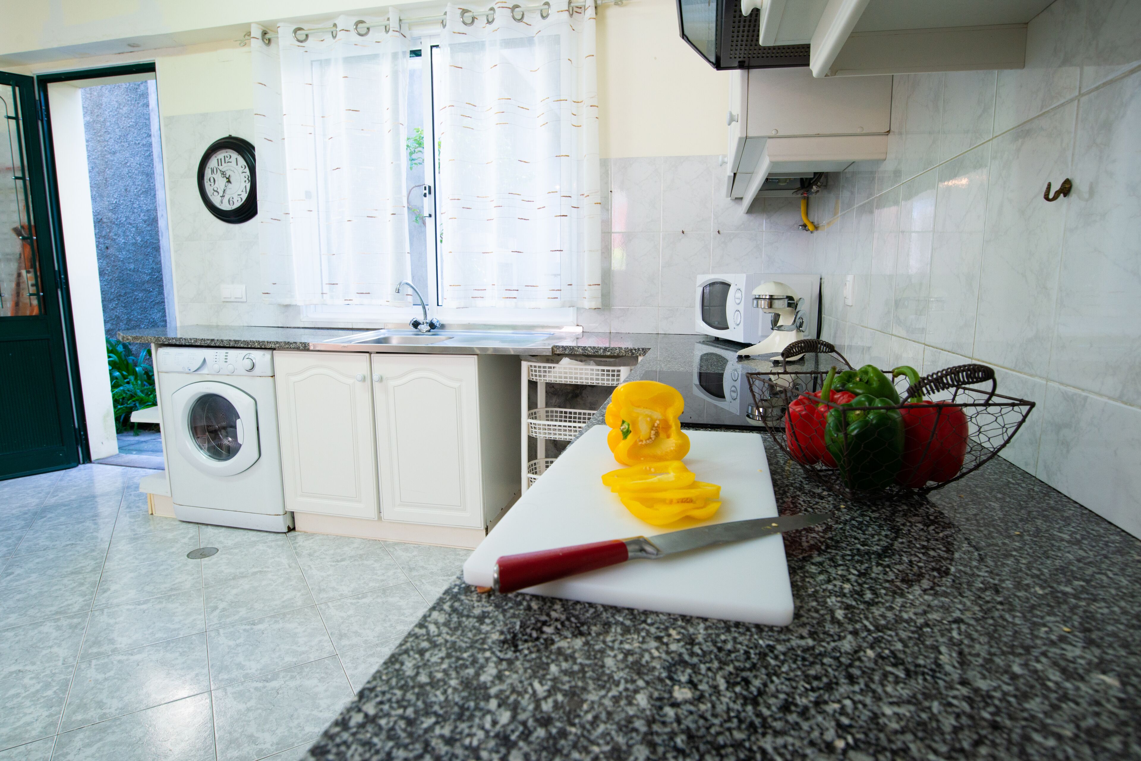 A kitchen with a washing machine, a sink, a microwave, and a cutting board with vegetables.