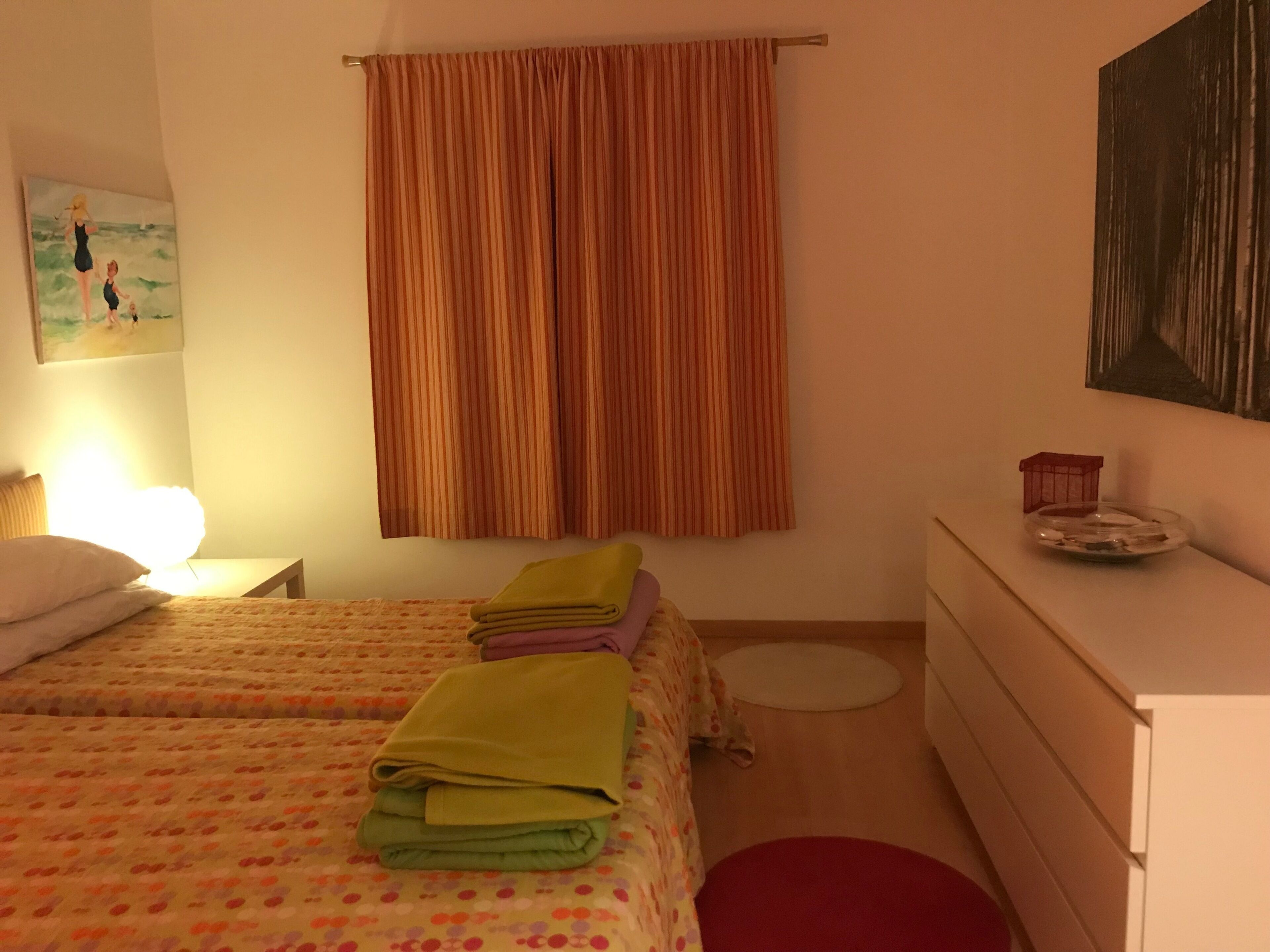 Habitación de hotel con dos camas, una mesita de noche con lámpara, cómoda y una alfombra roja, además de un cuadro en la pared.