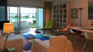 Un salon moderne avec une table en verre, deux fauteuils, un canapé, une bibliothèque et une vue sur une piscine et un immeuble à l’extérieur.