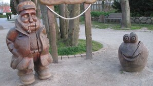 Zwei Holzskulpturen, eine wie ein Mann und die andere wie eine Eule, befinden sich in einer Parkanlage mit Bäumen und einer Bank im Hintergrund.