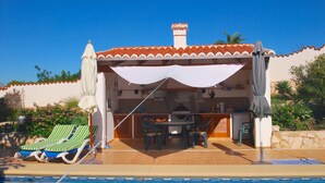 Uma área à beira da piscina com cozinha externa coberta, espreguiçadeiras e uma mesa de jantar.