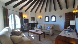 Uma sala de estar aconchegante com um sofá, uma mesa de centro e uma lareira. Há janelas com cortinas, uma televisão e um abajur.