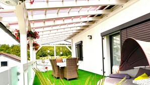 A covered outdoor area with a green artificial turf, wicker furniture, and a view of a house in the background. - Nestled between Rennsteig and the UNESCO Biosphere Reserve Thuringian Forest (Nahetal-Waldau)