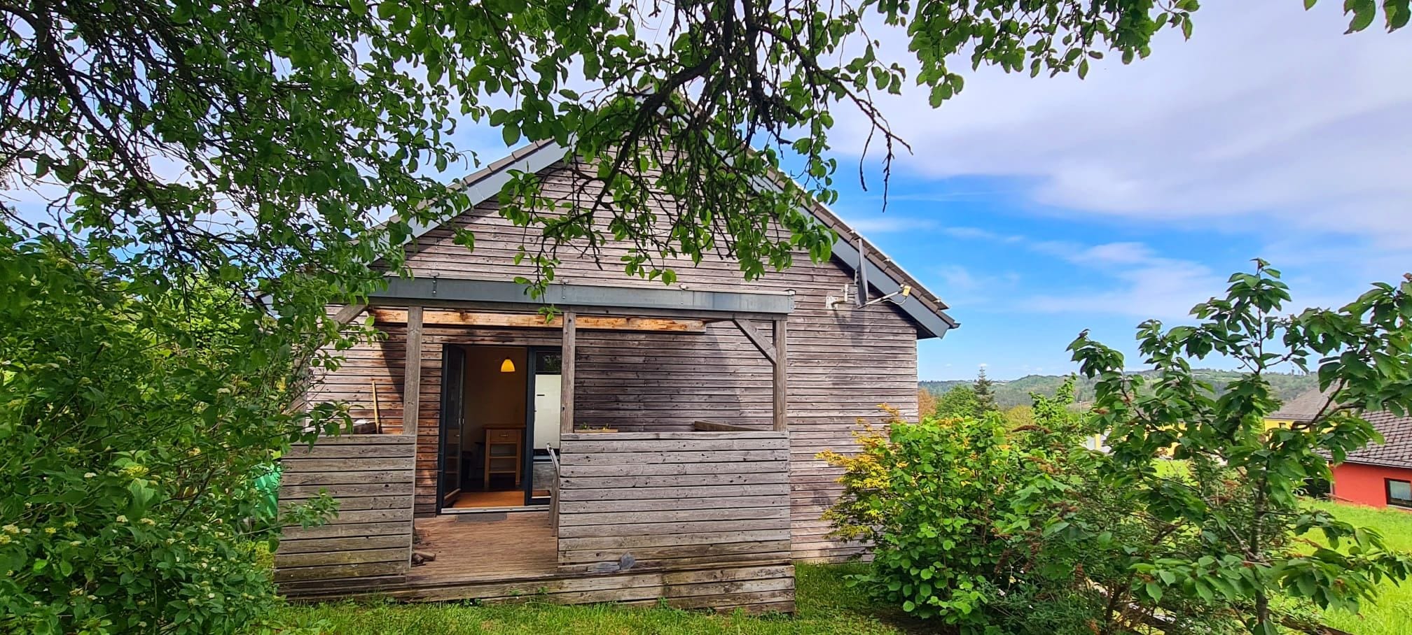 A rustic wooden cabin with a porch, surrounded by greenery and a clear sky.