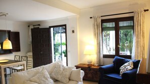 A living room with a white sofa, a blue armchair, a wooden side table with a lamp, and a view of greenery through a window with curtains.