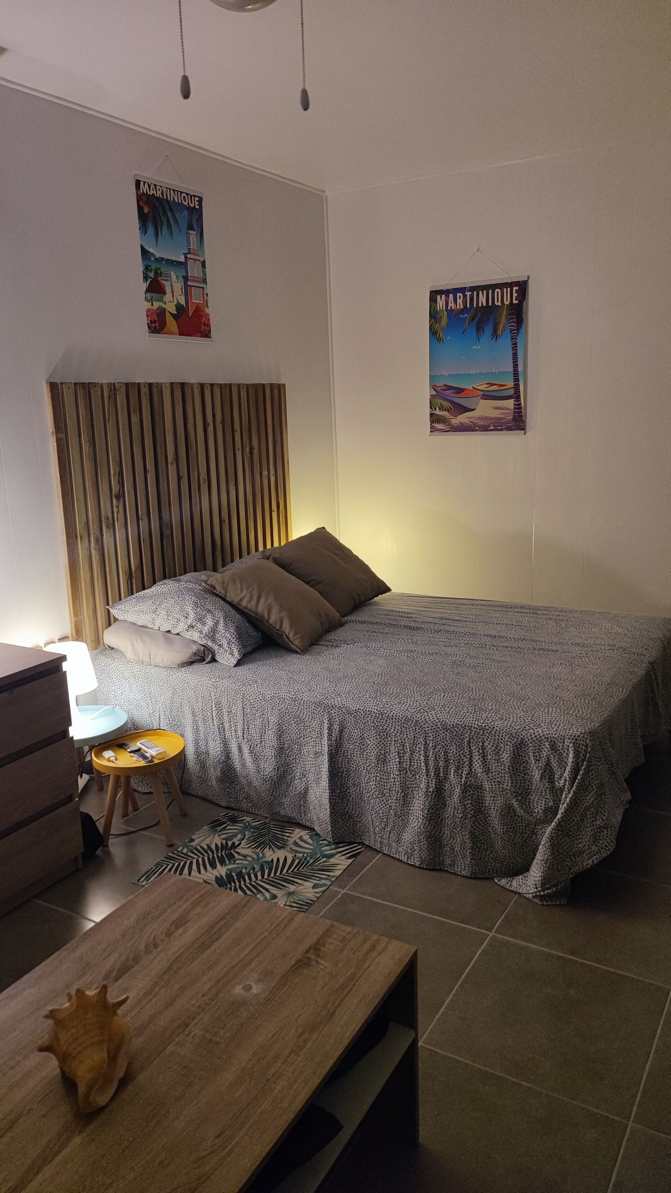 Une chambre à coucher avec un lit, une tête de lit en bois, une table de chevet, une lampe, une petite table ornée d’un coquillage et deux affiches accrochées au mur.