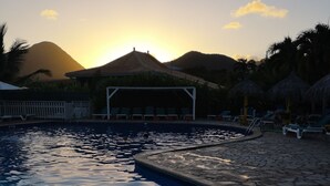 Uma piscina com espreguiçadeiras e uma estrutura com telhado de palha, cercada por palmeiras e montanhas ao fundo.