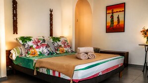 A neatly made bed with floral bedding and a striped blanket, a small bedside table with a lamp, and a painting of two figures on the wall. - Large villa **** beachfront with pool  (La Saline-Les-Bains)