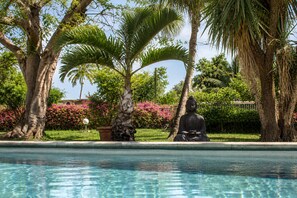Un espace piscine paisible, agrémenté d’une statue de Bouddha, entouré d’une végétation luxuriante et de plantes en fleurs.