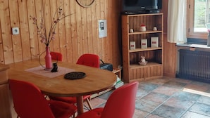 Une pièce aux murs lambrés, avec une table ronde en bois, des chaises rouges, un téléviseur posé sur une étagère en bois et une fenêtre avec des rideaux.