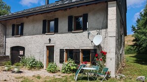 Une maison de campagne en pierre, dotée d’un toit en pente, de nombreuses fenêtres avec volets, d’une antenne parabolique et d’un espace salon extérieur.