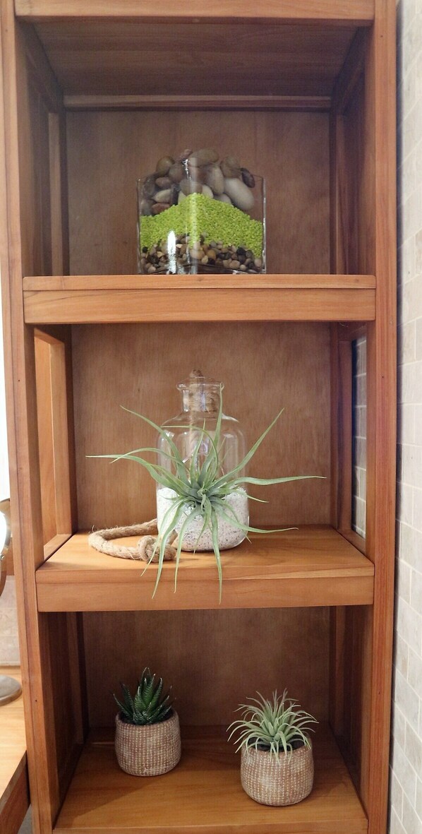 Una estantería de madera con tres repisas. La repisa superior sostiene un recipiente de vidrio con una planta verde y guijarros. La repisa central tiene un recipiente de vidrio transparente con una sola planta verde. La repisa inferior tiene dos macetas pequeñas.