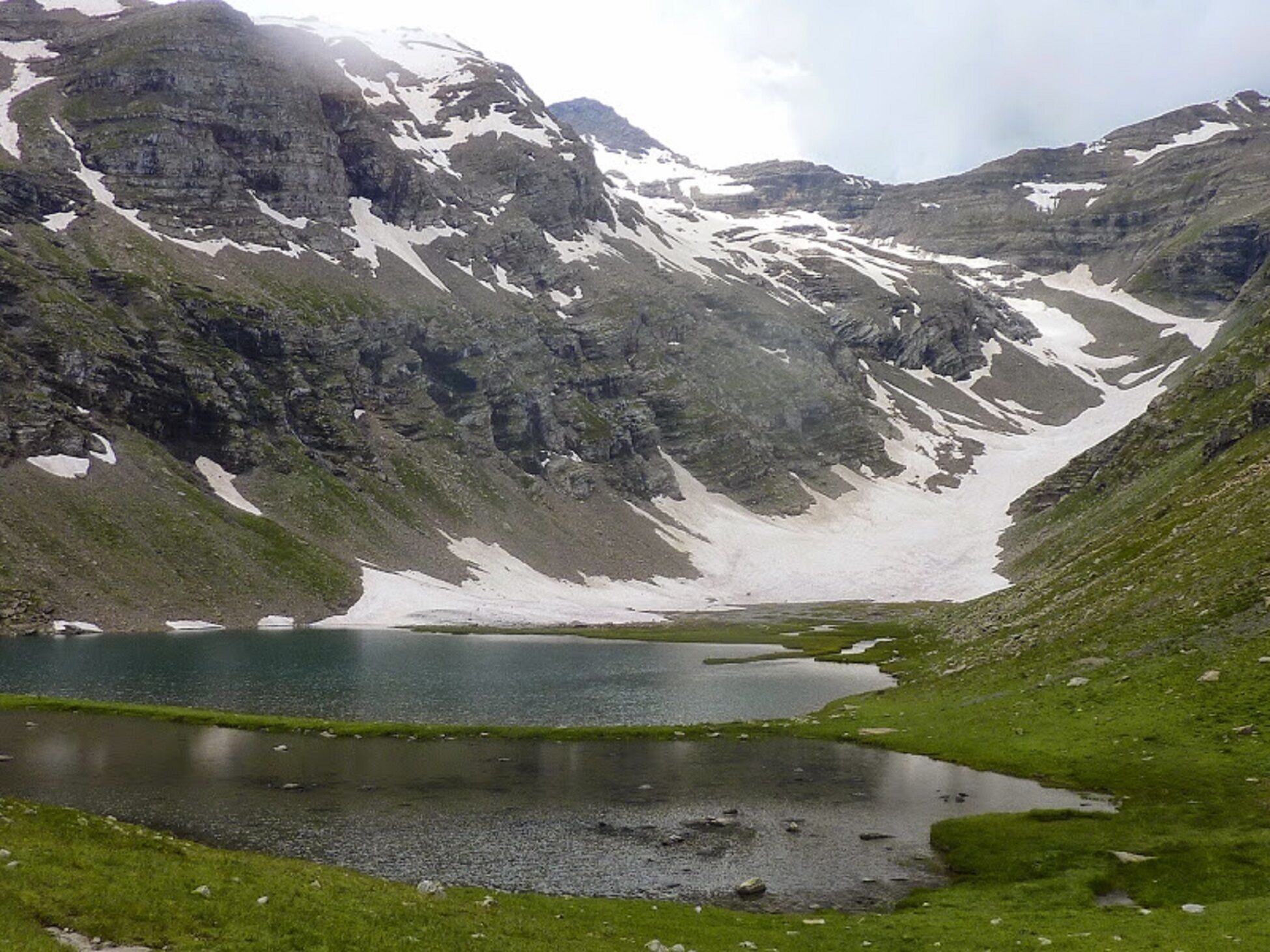 Paisagem montanhosa com um lago rodeado por terreno rochoso e algumas manchas de neve.