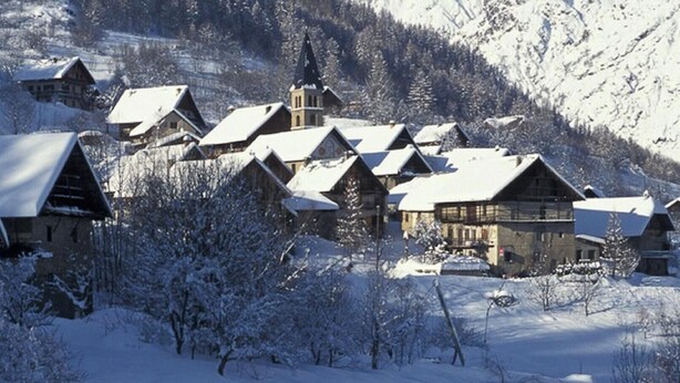 Ein traditionelles Chaletdorf aus Holz mit schneebedeckten Dächern und Bäumen, vor einer Bergkulisse gelegen.